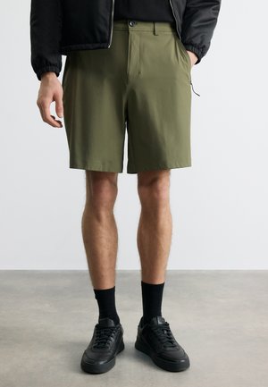 Bărbat purtând pantaloni scurți kaki de lungime până la genunchi, șosete negre și adidași negri, stând pe podea de beton în fața unui perete alb.