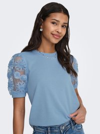 Jeune femme aux longs cheveux foncés portant un haut bleu clair avec des manches bouffantes en tulle brodées de motifs floraux, des boucles d'oreilles argentées et un jean en denim.