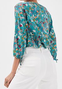 Blouse fleurie teal avec des manches bouffantes, des accents froncés et des détails à nouer aux poignets, associée à un pantalon blanc taille haute.