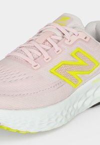 Ανοιχτό ροζ αθλητικό παπούτσι New Balance με κίτρινο λογότυπο, λευκή μαλακή σόλα και ροζ κορδόνια, σχεδιασμένο για άνεση και απόδοση.