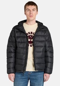 Man i en svart puffjacka över en ljusfärgad t-shirt med en logotyp, stående mot en enfärgad vit bakgrund.