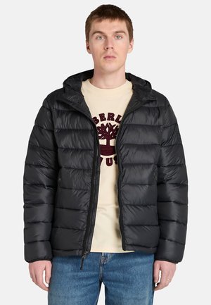 Man in een zwarte gewatteerde jas over een lichtgekleurde t-shirt met een logo, staande tegen een effen witte achtergrond.