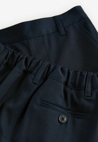 Dettaglio di pantaloni blu navy con cintura piatta, sezione elastica e tasca posteriore con bottone. Il materiale appare liscio con una sottile texture.