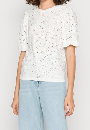 Femme portant un chemisier blanc à manches courtes brodées avec manches bouffantes et un jean bleu clair taille haute sur un fond uni.
