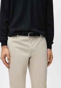 Pantaloni beige con una texture liscia, dotati di passanti per cintura e fibbia argentata. Abbinati a una camicia nera aderente a maniche lunghe.