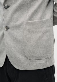 Ανοιχτό γκρι υφασμάτινο σακάκι με μεγάλη τσέπη επάνω και δύο σκούρα κουμπιά πάνω σε μαύρο παντελόνι, εμφανιζόμενο από τη μέση έως το μέσο του μηρού.