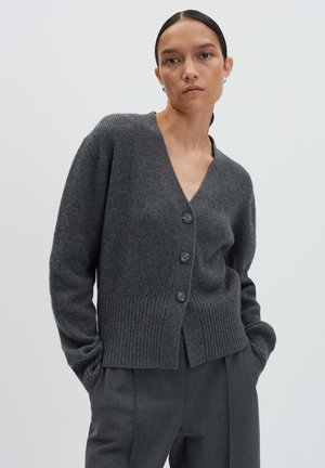Femme portant un cardigan boutonné gris foncé et un pantalon tailleur assorti, les mains dans les poches, sur un fond clair.