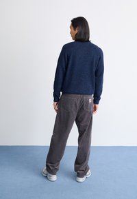 Granatowy sweter dzianinowy o teksturowanym wykończeniu, w połączeniu z luźnymi szarymi spodniami z kordurowej tkaniny z tylnymi kieszeniami. Model stoi na tle niebieskiego dywanu.