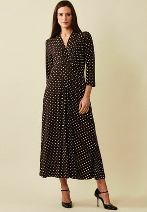 Modell som bär en långärmad klänning med bruna prickar och svarta klackar, stående mot en ljus beige bakgrund, med ett neutralt uttryck.