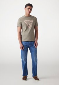 Khaki kr_shortām t-kreklu ar trīsstūrainu rozā un baltu grafisko logotipu centrā, apvienots ar gaiši ziliem džinsiem un brūnām kurpēm.