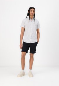 Camicia a maniche corte bianca con bottoni e un motivo sottile, abbinata a pantaloni corti neri. Il modello indossa scarpe da ginnastica beige e ha i capelli intrecciati.
