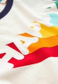 Witte stoffen t-shirt met meerkleurige "HAPPY" tekst in vetgedrukte, blokletters. Kleuren zijn onder andere rood, oranje, geel en teal accenten.