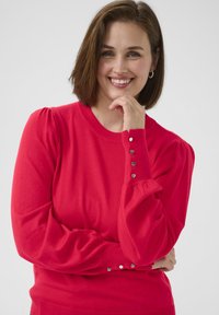 Red knit sweater featuring long puffed sleeves, ribbed crew neckline, and decorative silver buttons on the cuffs. Smooth texture.