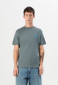 Ung mand iført en afdæmpet grøn t-shirt med en blå grafik af en skrigende hånd på brystet, parret med blå denimbukser, stående neutralt.