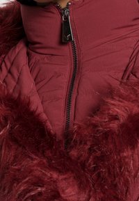 Gros plan sur une veste matelassée bordeaux fermée par une fermeture éclair avec un tire-zip argenté et un col en fausse fourrure bordeaux.