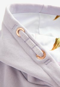 Tessuto lilla chiaro con una texture liscia, dotato di un occhiello in oro rosa e una tracolla testurizzata con dettagli stampati. Fodera floreale visibile.