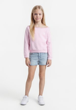 Meisje met lang blond haar staat frontaal, draagt een lichtroze off-shoulder sweatshirt, lichte denim shorts en witte sneakers.