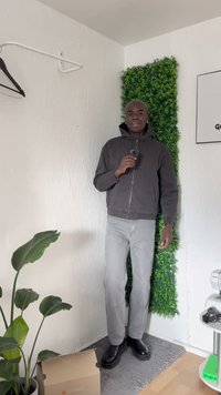 Czarna bluza z kapturem na zamek, szare dżinsy i czarne botki za kostkę. Tło przedstawia zieloną roślinę i sztuczną ścianę z trawy.