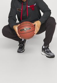 Sudadera gris con cordones rojos y un logo de los Chicago Bulls, pantalones deportivos negros, sosteniendo un balón de baloncesto Spalding, usando zapatillas deportivas negras y grises.