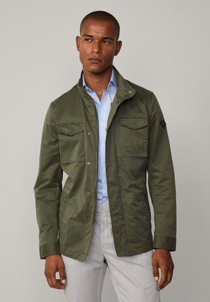 Olijfgroene jas met een rits aan de voorkant, drukknopen, twee borstzakken en een logo-accent op de mouw; lichte stof met een gladde textuur.