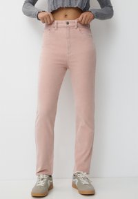 Pantalon taille haute en velours côtelé rose clair, coupe slim, avec poches avant et fermeture par boutons. Porté avec des baskets grises.