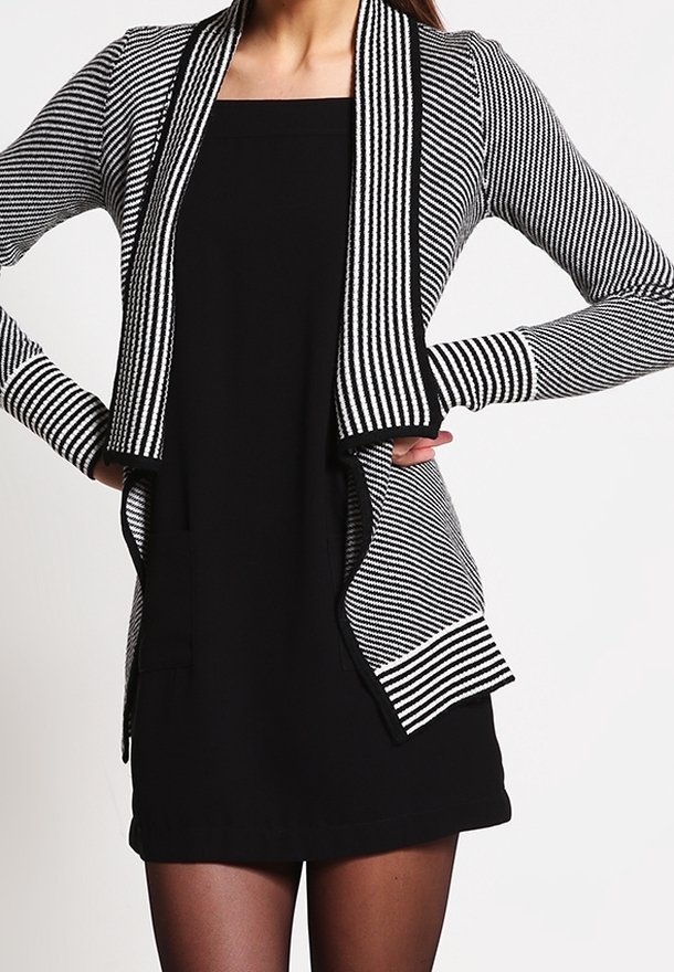 Femme portant une robe noire et un cardigan rayé noir et blanc, mains sur les hanches, sur un fond uni.