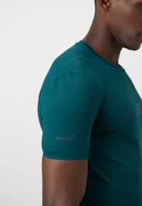Teal athletic shirt with a fitted design. Features short sleeves, a round neckline, and a "VIGOR7" logo on the left sleeve. Smooth texture.