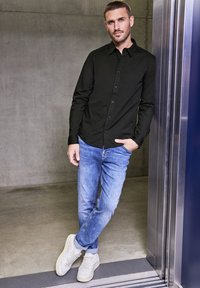 Zwarte langemouwen knooptrui, lichtblauwe jeans en witte sneakers; staand tegen een lift met een betonnen achtergrond.