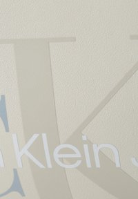 Detailní záběr na texturovaný béžový povrch s překrývajícími se světle šedými a bílými písmeny loga Calvin Klein.