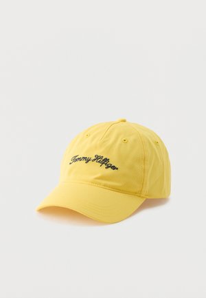 Șapcă galbenă de baseball cu logo „Tommy Hilfiger” negru, scris de mână brodat în față, prezentată pe fundal alb simplu.