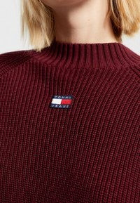 Burgundy knit sweater with a ribbed neckline, featuring a blue and red branded patch labeled "TOMMY JEANS" on the front.
