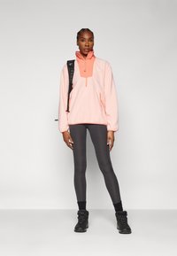 Vaaleanpunainen fleece-pusero, jossa on puolivetoketju ja läpällinen tasku, päällä tummanharmaat legginssit ja mustat nauhaketjulliset saappaat.