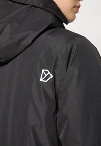 Crna vodootporna jakna s kapuljačom, glatke teksture i bijelim geometrijskim logotipom na lijevom ramenu.