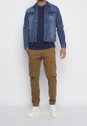 Jeansjacke mit Fronttaschen, blau-weiß gestreiftes T-Shirt, beigefarbene Cargohosen mit Seitentaschen und weiße Turnschuhe.