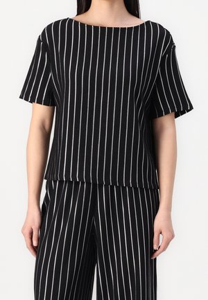 Kobieta w czarnej koszulce z krótkim rękawem i pasujących szerokich spodniach z pionowymi białymi prążkami na jednolitym tle.