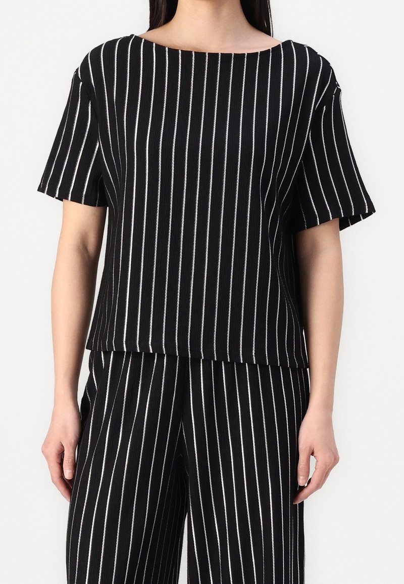 Femme portant un haut noir à manches courtes et un pantalon large assorti avec des fines rayures blanches verticales sur un fond uni.