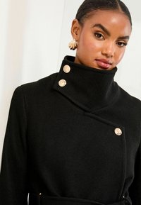 Melna vilnas mētelis ar augstu apkakli, kam piemīt metāla pogu akcents. Dizainā ir ietverta diagonāla aizdare un teksturēta auduma izmantošana.