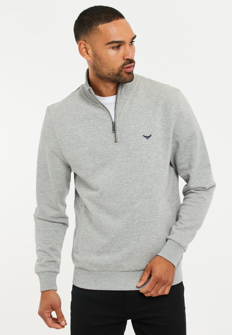 Homme portant un sweat-shirt gris clair avec fermeture éclair quart de zip et petit logo brodé, debout et regardant vers sa gauche sur un fond uni.