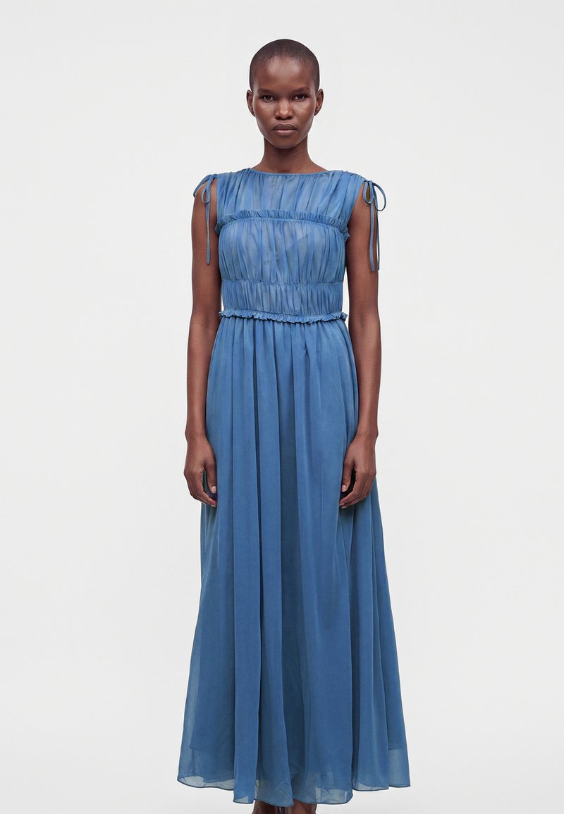 Femme portant une robe bleue sans manches longue jusqu'au sol avec un corsage froncé et des bretelles nouées, debout devant un fond blanc uni.