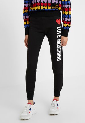 Personne portant un pantalon de survêtement noir avec un cœur rouge et le texte "LOVE MOSCHINO" sur la jambe, un haut coloré à motifs de cœurs et des baskets blanches.