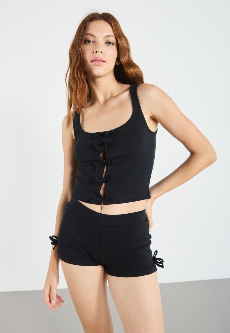 Melnā crop top ar pogu un loku akcentiem un kvadrātveida dekoltē, apvienots ar atbilstošām melnām šortiem ar aukliņu detaļām sānā.