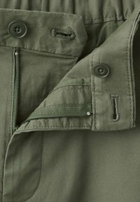 Gros plan sur la fermeture éclair ouverte et la fermeture à bouton d'un pantalon vert montrant les boutonnières et les détails de couture.