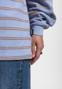 Gestreepte lange mouw shirt in lichtblauw en bruin, met een gestructureerde stof. Een label met "DRE DENIM" is zichtbaar aan de zoom. Hand rustend op denim.