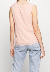 Top sin mangas de color rosa claro, hecho de una tela suave, con un escote redondo y un dobladillo trasero un poco más largo, combinado con jeans azul claro.