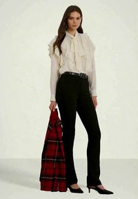 Moteris, vilkinti kreminės spalvos raukšlėtą bluzą ir juodas kelnes, laiko raudoną languotą švarką, stovinti su juodais aštriais bateliais ant švies background.