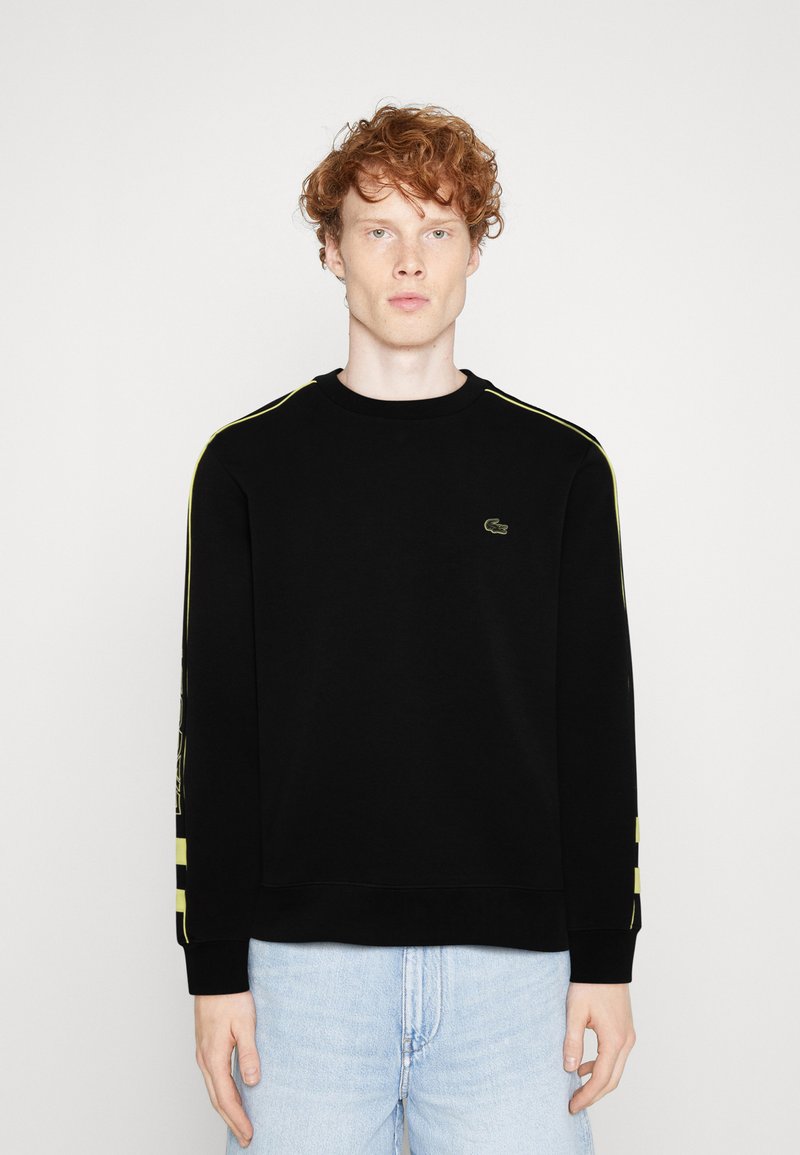 Sweatshirt noir avec des manches longues, un col rond, un logo vert sur la poitrine et des bandes jaunes sur les côtés pour l'accent. Design décontracté.