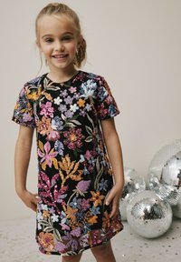 Schwarzes Kleid, verziert mit buntem Blumenstick und Pailletten. Kurze Ärmel, runder Ausschnitt und knielanges Design. Silberne Discokugeln im Hintergrund.