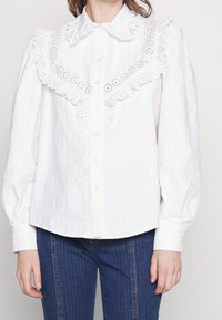 Camicia di cotone bianco con colletto in pizzo arricciato, bottoni frontali e righe a trama. Presenta maniche lunghe e un orlo arrotondato.