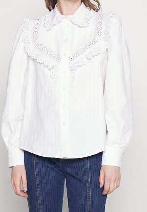 White cotton blouse with a ruffled lace collar, front buttons, and textured stripes. Features long sleeves and a rounded hem.