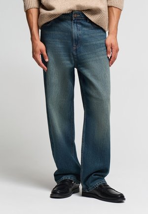 Uomo con jeans blu larghi con dettagli sbiaditi, maglione beige e mocassini neri in pelle, in piedi davanti a uno sfondo semplice.